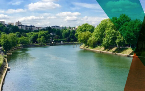 Paysage urbain naturel avec un fleuve entouré d'arbres, avec des bâtiments visibles au loin.