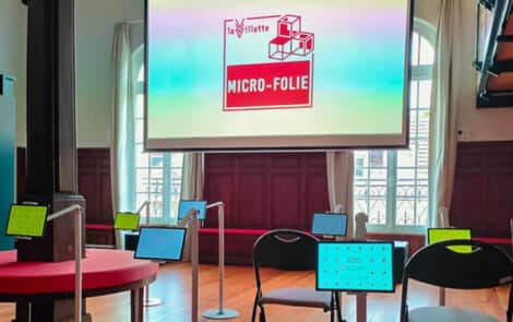 Salle d'exposition avec écrans tactiles et table ronde rouge, logo Micro-Folie de La Villette sur grand écran, chaises noires autour.